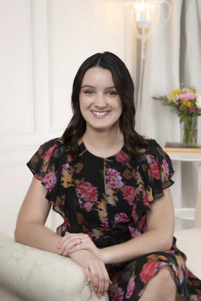 A woman with dark hair in loose waves, wearing a black dress with a colorful floral pattern, sits smiling on a light sofa—perhaps a wedding planner taking a break from wedding planning. Behind her, there is a vase of flowers and a lamp on a table. - Jodi O Photography