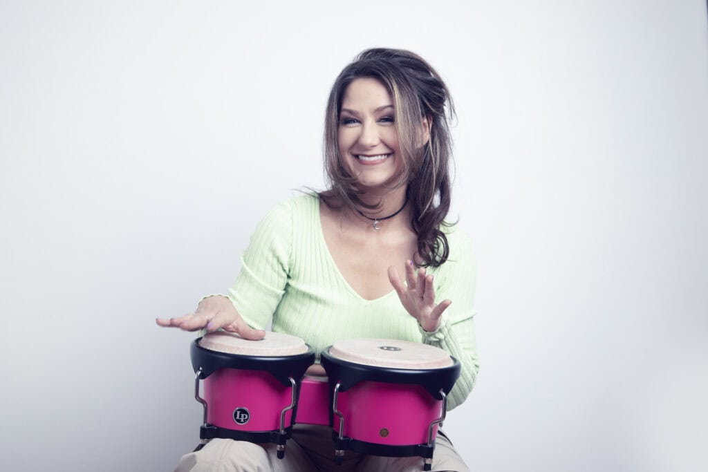 A smiling woman with long brown hair plays two pink bongos in a studio. She wears a light green top and sits against a plain, light background. - Jodi O Photography