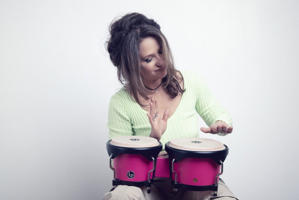 A woman with long brown hair wearing a light green top plays two bright pink bongo drums in a music studio, looking down at the drums with a slight smile against a plain white background. - Jodi O Photography