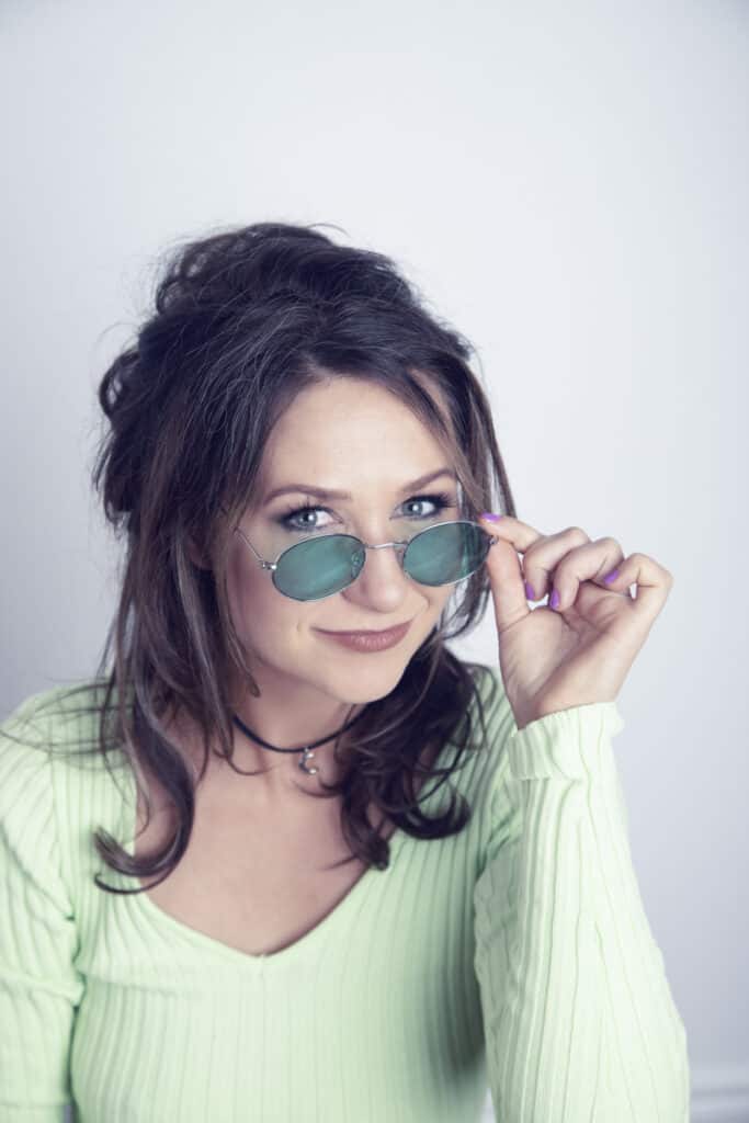A woman with long brown hair wearing a light green ribbed top and round green sunglasses looks at the camera, smiling slightly and lifting her glasses with one hand in a music studio, radiating Rock Star Energy. - Jodi O Photography