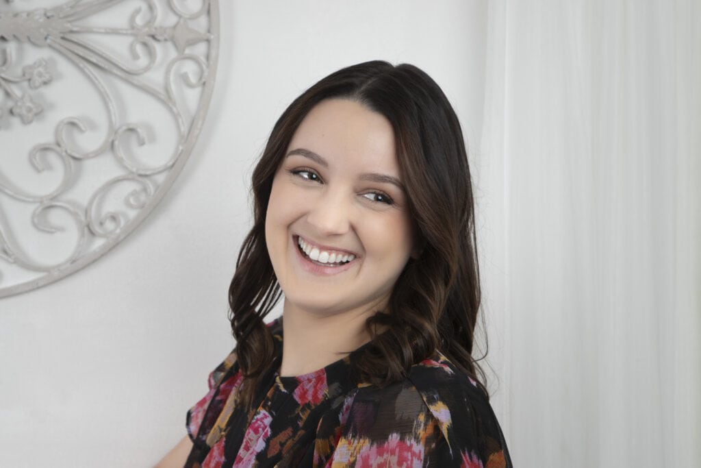 A young woman with long, wavy brown hair smiles brightly while looking slightly to the side. Wearing a colorful, patterned top, she stands in front of a white wall with decorative metalwork, radiating confidence as she pursues her dream of building a brand. - Jodi O Photography