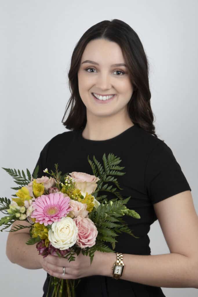A woman with long brown hair, wearing a black short-sleeve top and a gold wristwatch, smiles while holding a colorful bouquet—perfect inspiration for wedding planning—against a plain light background. - Jodi O Photography