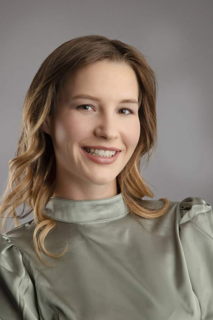 A woman with light brown hair and a light green blouse smiles at the camera, creating a fresh modern business image against a soft gray background. - Jodi O Photography