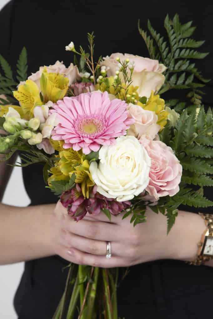 A person in a black outfit, possibly a wedding planner, holds a bouquet of pink gerbera, white and pale pink roses, yellow alstroemerias, and greenery. A gold watch and ring are visible on their left hand. - Jodi O Photography