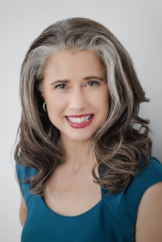 Confident and authentic, a smiling woman with long, wavy salt-and-pepper hair wears a teal dress and silver hoop earrings, posing against a plain light background. - Jodi O Photography