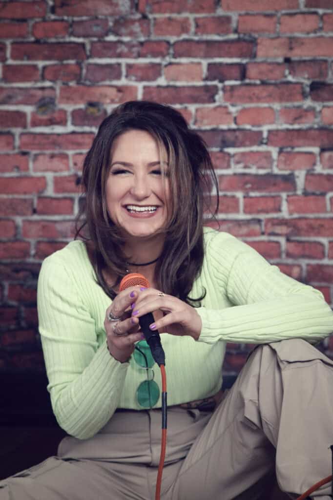 A woman with long brown hair, wearing a light green ribbed top and beige pants, sits against a brick wall, smiling and holding an orange microphone as if she’s ready to record in a music studio. Green sunglasses hang from her waist. - Jodi O Photography