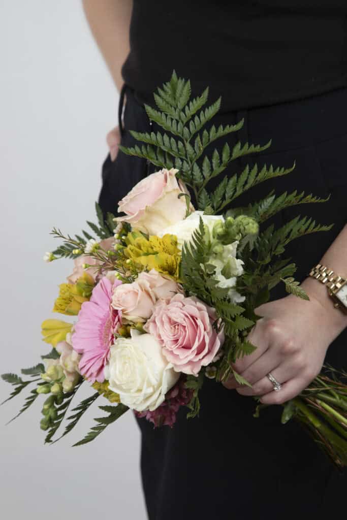 A person in black clothes, perhaps a wedding planner building a brand, holds a colorful bouquet of flowers—roses and ferns—in one hand, with a gold watch and ring on their left. The background is plain and light-colored. - Jodi O Photography