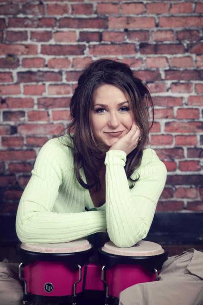 A woman with long brown hair and a light green top leans on her hand, smiling at the camera in a music studio. She rests her arm on two pink bongo drums, with a brick wall background behind her. - Jodi O Photography