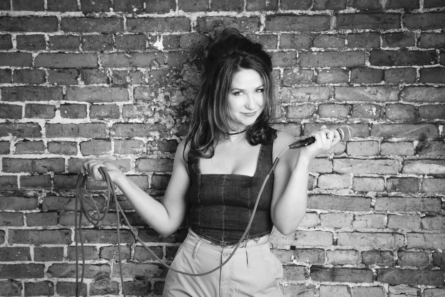 A woman stands in front of a brick wall holding a microphone and its cable, as if ready for a studio session. She wears a sleeveless top and high-waisted pants, smiling slightly. The black and white image exudes subtle Rock Star Energy. - Jodi O Photography