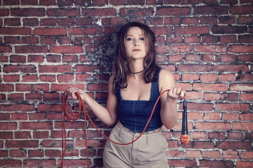 A woman stands against a brick wall, holding a coiled microphone cable and a microphone—ready for her next recording. Wearing a sleeveless top and light-colored pants, she exudes confidence like she's in her own music studio. - Jodi O Photography