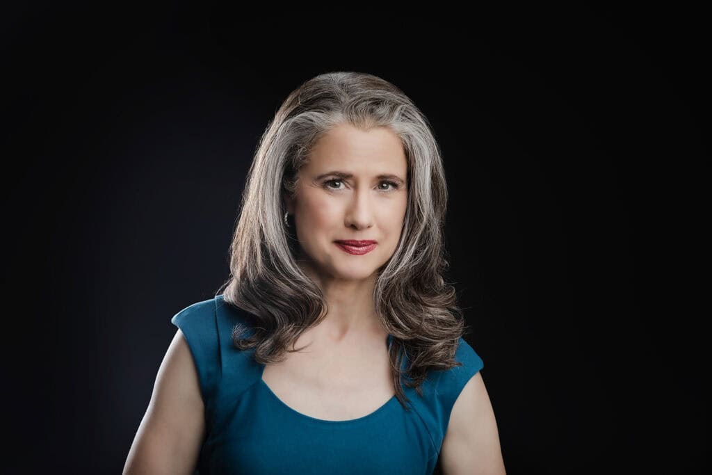 A confident woman with long, wavy gray hair and red lipstick, wearing a teal dress, poses against a black background, looking authentic and self-assured at the camera. - Jodi O Photography