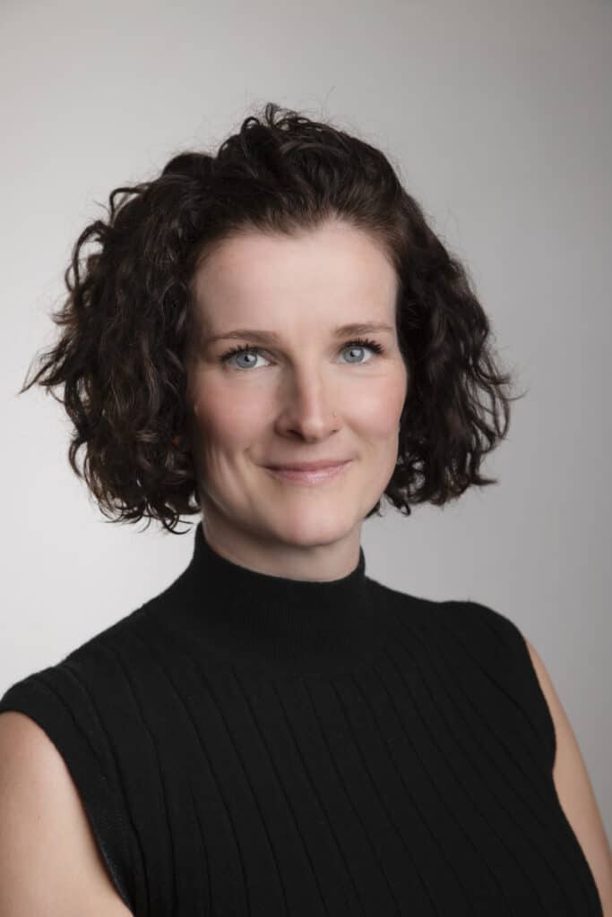 A woman with short, curly brown hair and blue eyes gives a warm, slight smile. She wears a sleeveless black turtleneck top and is posed against a plain, light gray background for a natural, professional look. - Jodi O Photography