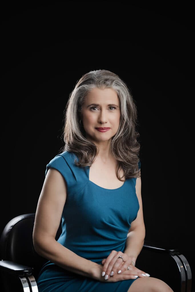 A confident woman with long, wavy gray hair sits on a black chair against a dark background. She wears a teal dress and has her hands folded on her lap, looking calmly and authentically at the camera. - Jodi O Photography