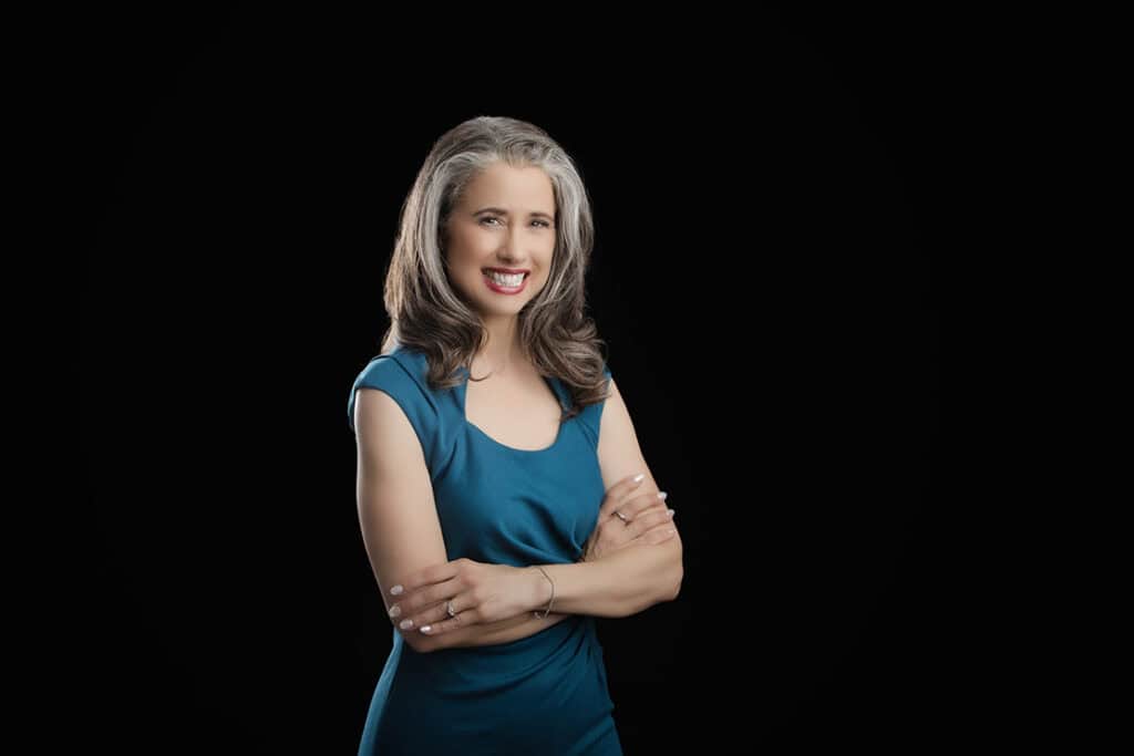 A confident, smiling woman with wavy gray hair wears a teal dress and stands with arms crossed against a solid black background. - Jodi O Photography