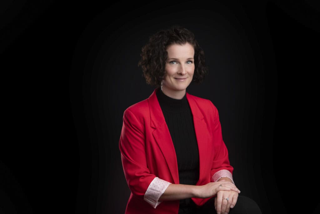 A professional woman with natural, curly brown hair wears a bright red blazer over a black top, sitting against a black background. She smiles gently, her hands resting warmly on her knee. - Jodi O Photography