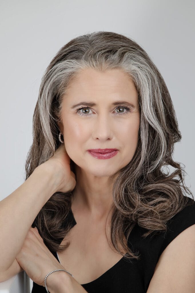 A professional woman with long, wavy gray and brown hair wears a black top and touches her neck. With light makeup, red lipstick, and an authentic air, she looks confidently at the camera against a light background. - Jodi O Photography
