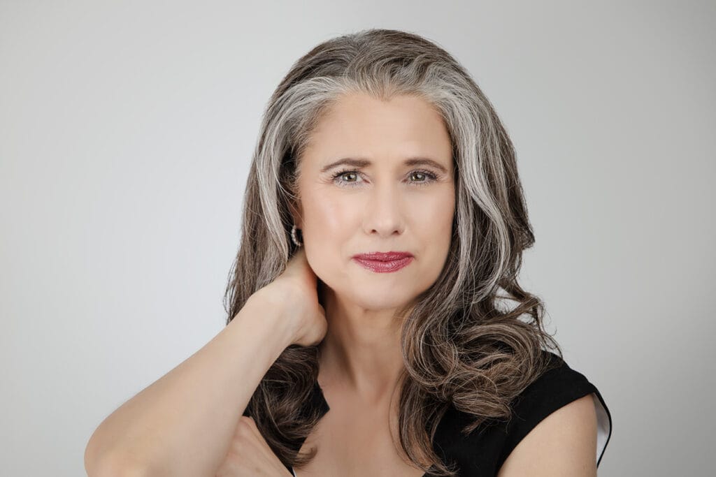 A confident woman with long, wavy gray and brown hair poses with her hand behind her neck, wearing a black top and subtle makeup, projecting a professional yet authentic presence against a plain light gray background. - Jodi O Photography