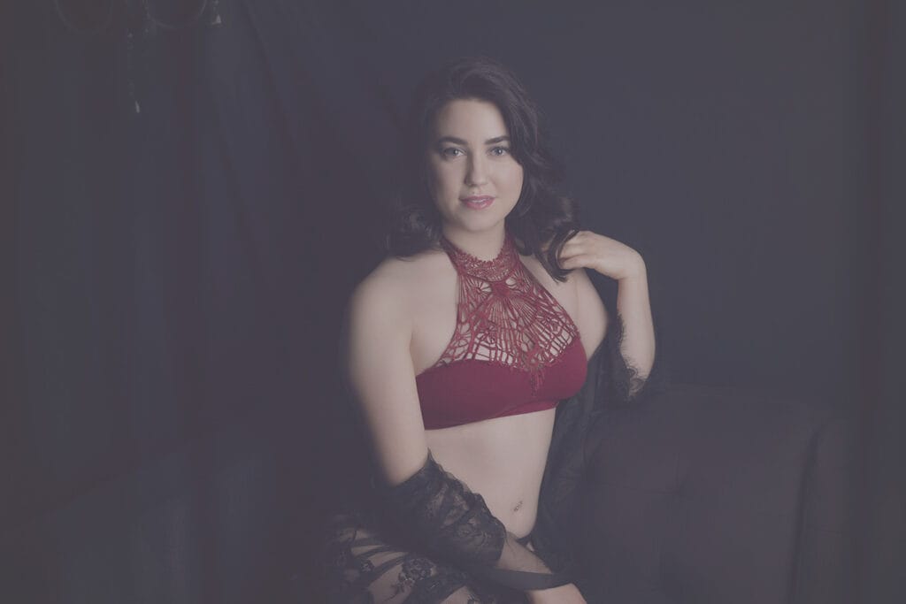 A woman with dark hair sits on a dark couch, wearing a burgundy halter bralette and a black lace shawl, posing against a dark backdrop and smiling softly at the camera. - Jodi O Photography