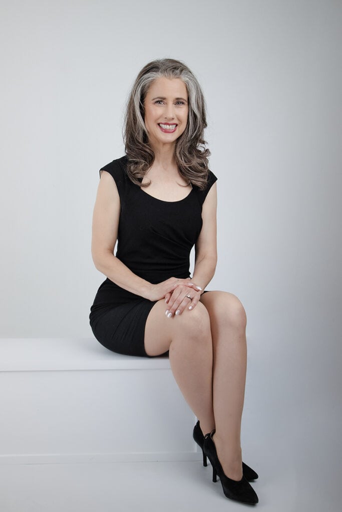 A confident woman with long gray hair wearing a short black dress and black heels sits on a white bench, smiling authentically at the camera against a plain white background. - Jodi O Photography