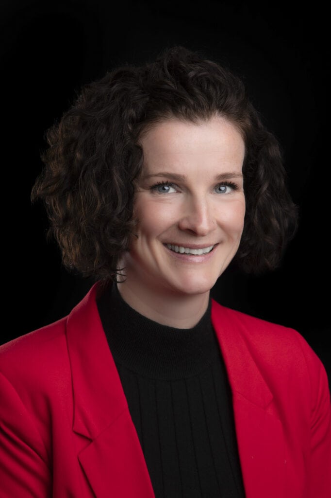 A professional woman with short, curly brown hair wearing a bright red blazer and black top, smiling warmly against a solid black background for a natural yet polished look. - Jodi O Photography