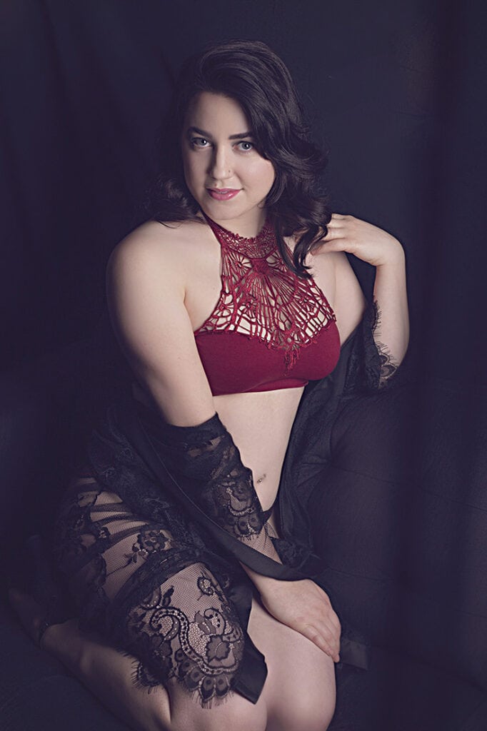 A woman with wavy dark hair sits on a dark surface, wearing a red lace halter bralette and a sheer black lace robe, looking at the camera with a slight smile. The background is dark and soft. - Jodi O Photography