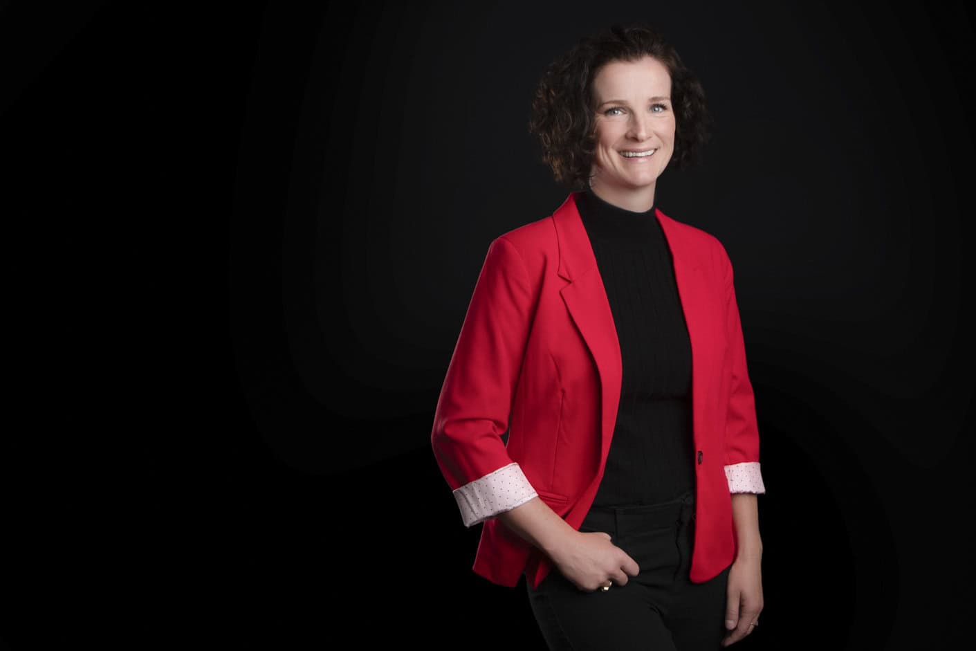 A professional with curly brown hair, wearing a bright red blazer over a black turtleneck and black pants, stands smiling warmly against a plain black background. - Jodi O Photography
