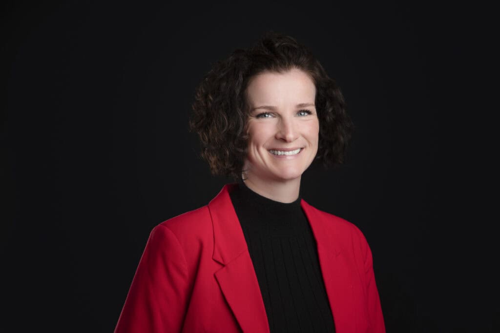 A warm, professional individual with short curly brown hair smiles while wearing a bright red blazer over a black top, posed against a solid black background. - Jodi O Photography