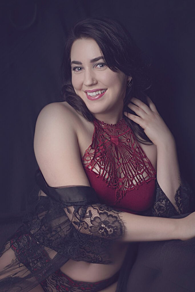 A woman with dark hair smiles while posing in a red lace halter lingerie set and a sheer black lace robe, against a dark background. - Jodi O Photography