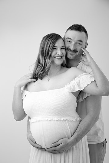 A smiling pregnant woman in a maternity off-shoulder dress stands in front of a man who hugs her from behind, both looking happy and affectionate, against a plain background. - Jodi O Photography