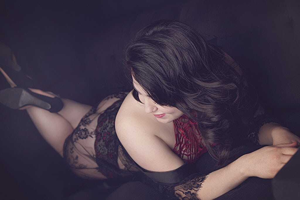 A woman with long dark hair lounges on a dark surface, wearing a sheer lace outfit with red accents. She lies on her stomach, smiling softly and looking downward, one arm bent and the other resting forward. - Jodi O Photography