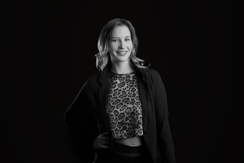 Black and white portrait of a woman smiling, wearing a leopard print crop top and blazer with one hand on her hip, standing against a dark background—a striking modern business image. - Jodi O Photography