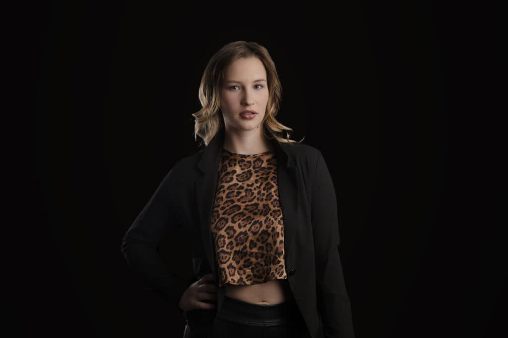 A woman with wavy, shoulder-length hair stands against a black background, wearing a black jacket over a leopard-print crop top—a striking modern business image, with one hand on her hip and a neutral expression. - Jodi O Photography