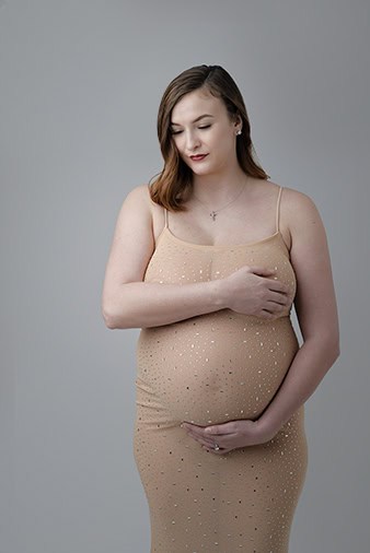 A pregnant woman in a sparkly, fitted beige dress stands against a plain gray background. Embracing her belly in a loving maternity pose, she gently covers her chest and gazes down with a calm expression—perfect for a maternity full post. - Jodi O Photography