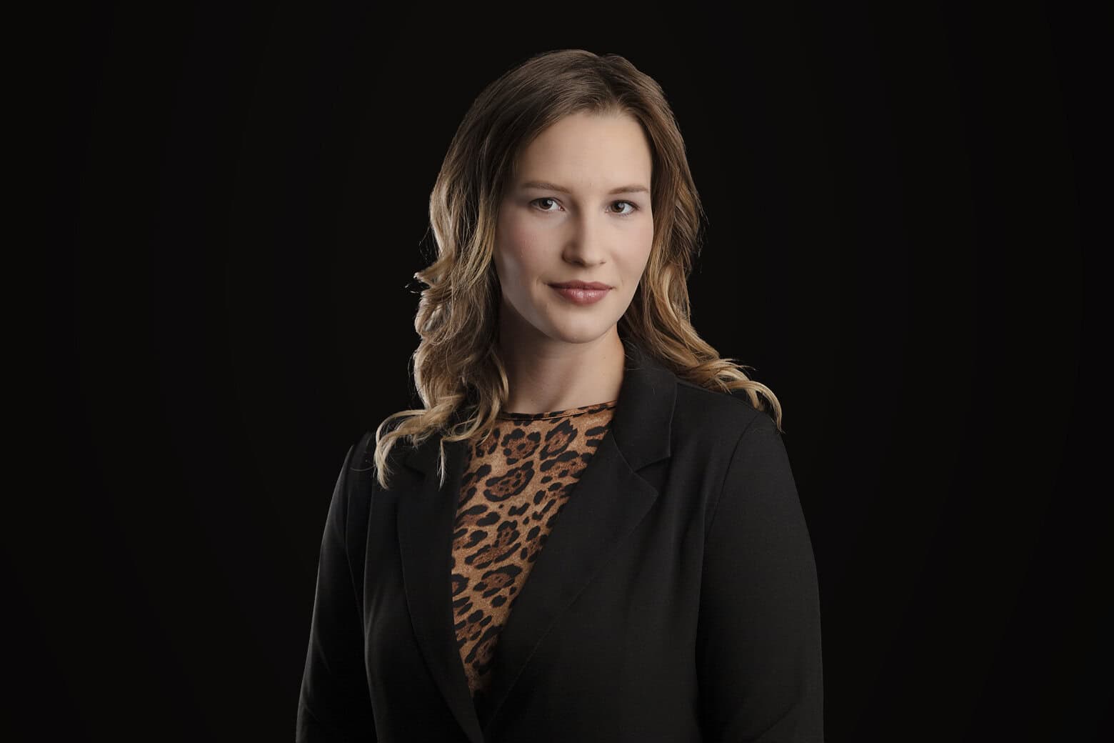 A woman with wavy, shoulder-length hair wearing a black blazer over a leopard-print top poses confidently against a plain black background, projecting a modern business image. - Jodi O Photography