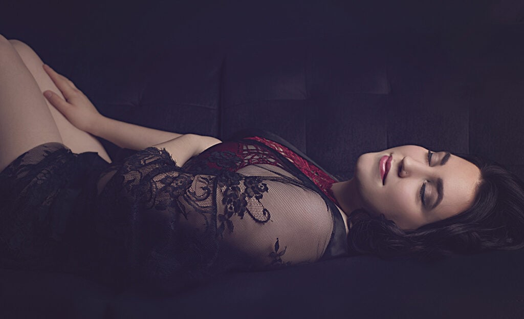 A woman with dark hair reclines on a dark surface, wearing lace and a red garment. Her eyes are closed and she has a peaceful expression, with one hand resting on her arm and the other by her side. - Jodi O Photography