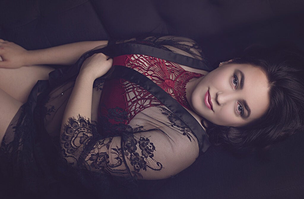 A woman with dark hair reclines on a dark surface, wearing a sheer black lace robe over a red lace bodysuit. She gazes at the camera with a soft expression, her hand resting near her shoulder. - Jodi O Photography