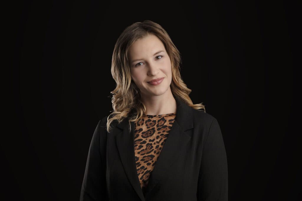 A woman with wavy light brown hair, wearing a black blazer over a leopard print top, stands against a solid black background, offering a slight smile—a confident modern business image. - Jodi O Photography