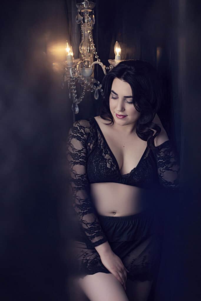 A woman in black lace lingerie poses softly against a dark background, illuminated by a chandelier. She looks down with a gentle expression, her dark hair styled in loose waves. - Jodi O Photography