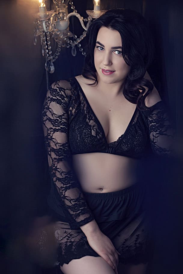 A woman with dark hair poses indoors wearing a lacy black bralette and sheer black robe, leaning against a dark wall near a lit chandelier, looking at the camera with a relaxed expression. - Jodi O Photography