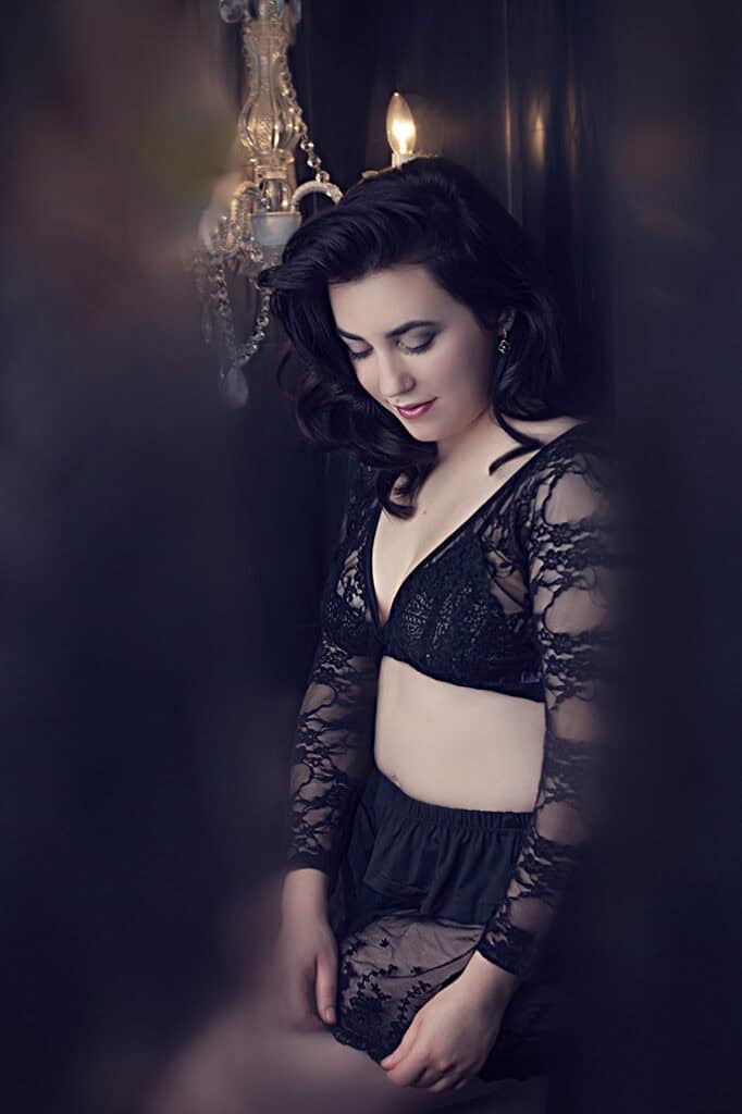 A woman in a sheer, black lace lingerie set stands with her head slightly bowed, softly lit by a chandelier in a dark, elegant room with a dreamy, intimate ambiance. - Jodi O Photography