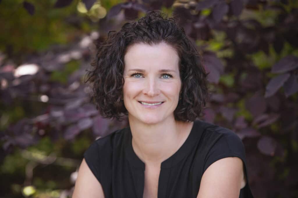 A woman with natural, curly dark hair and blue eyes smiles warmly at the camera, wearing a black top. She is outdoors with professional-looking, leafy purple and green foliage in the background. - Jodi O Photography