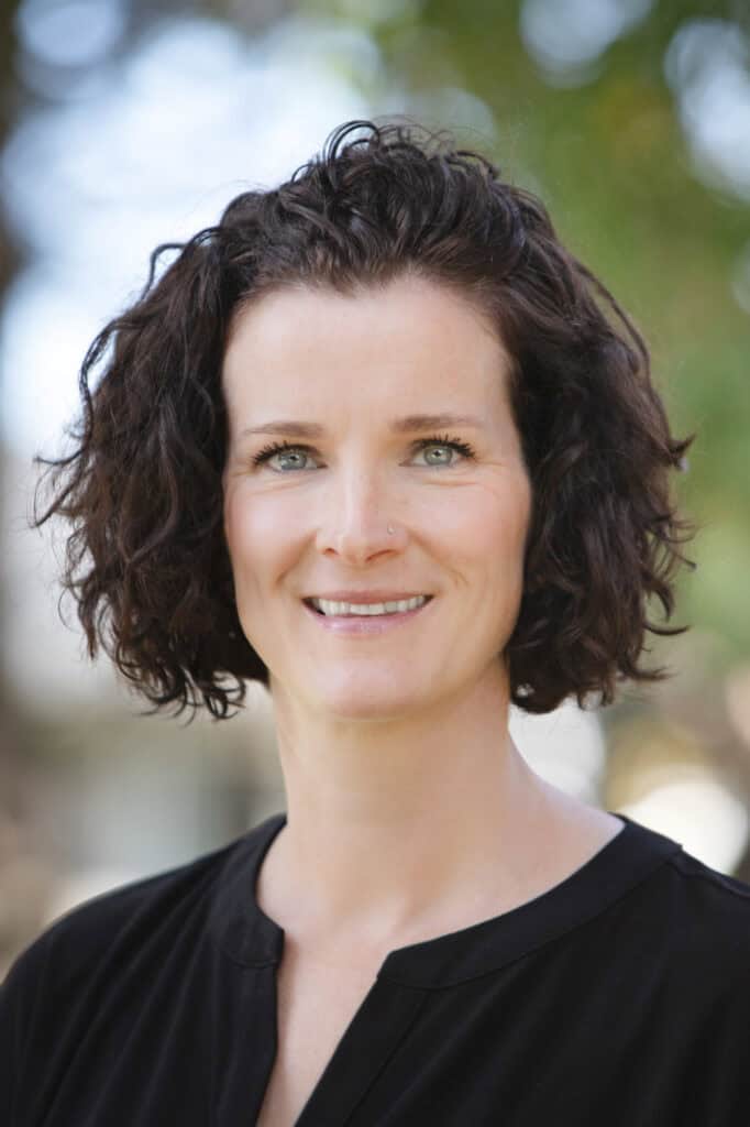 A woman with short, curly brown hair and blue eyes gives a warm smile to the camera. She is wearing a black top and is outdoors, with green trees and natural light softly blurred in the background. - Jodi O Photography