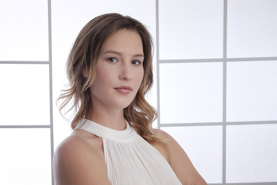 A woman with light brown hair and fair skin, wearing a sleeveless white top, stands with arms crossed in front of a bright, grid-patterned background, projecting a confident and modern business image. - Jodi O Photography