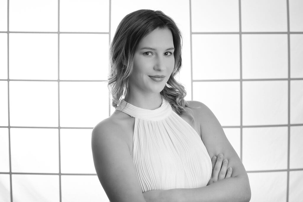 A woman with wavy hair in a sleeveless, high-neck white dress stands with arms crossed, smiling slightly. The black-and-white photo's grid background creates a modern business image perfect for business branding. - Jodi O Photography