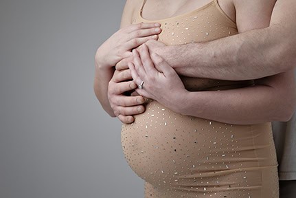 A pregnant person in a beige, sparkly dress is gently embraced from behind by another, with both pairs of hands resting on the baby bump—a tender moment perfect for a maternity blog. The pregnant person is wearing a ring. - Jodi O Photography