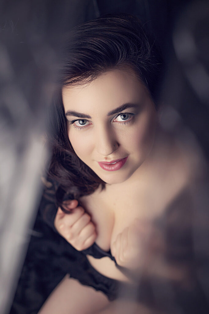 A woman with wavy dark hair and light eyes looks up at the camera, holding the straps of her black lingerie with both hands. The image has a soft, intimate feel and is slightly blurred around the edges. - Jodi O Photography