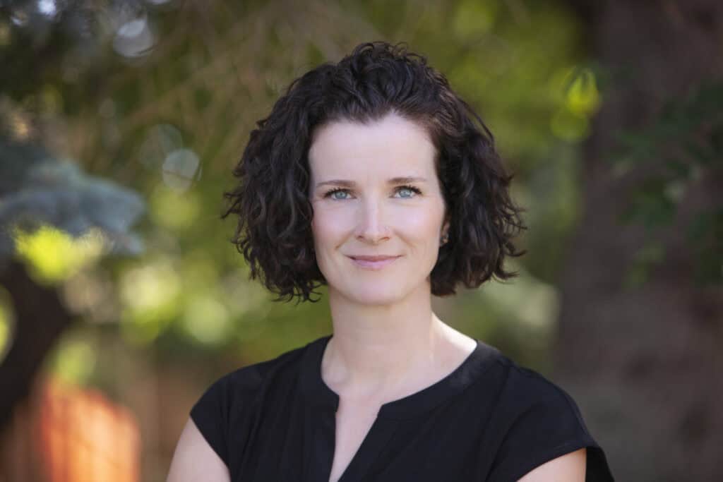 A warm, professional woman with short, curly dark hair and blue eyes is smiling gently, wearing a black blouse, standing outdoors with natural greenery and sunlight softly blurred in the background. - Jodi O Photography