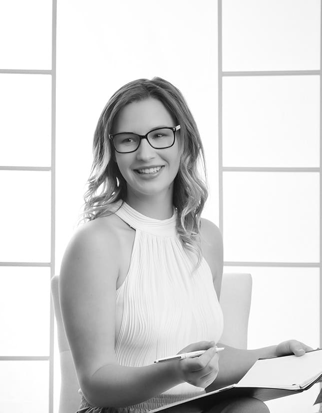 A woman with long hair and glasses, wearing a sleeveless white top, sits and smiles while holding a pen and notebook, capturing a modern business image against a bright, grid-patterned background. - Jodi O Photography