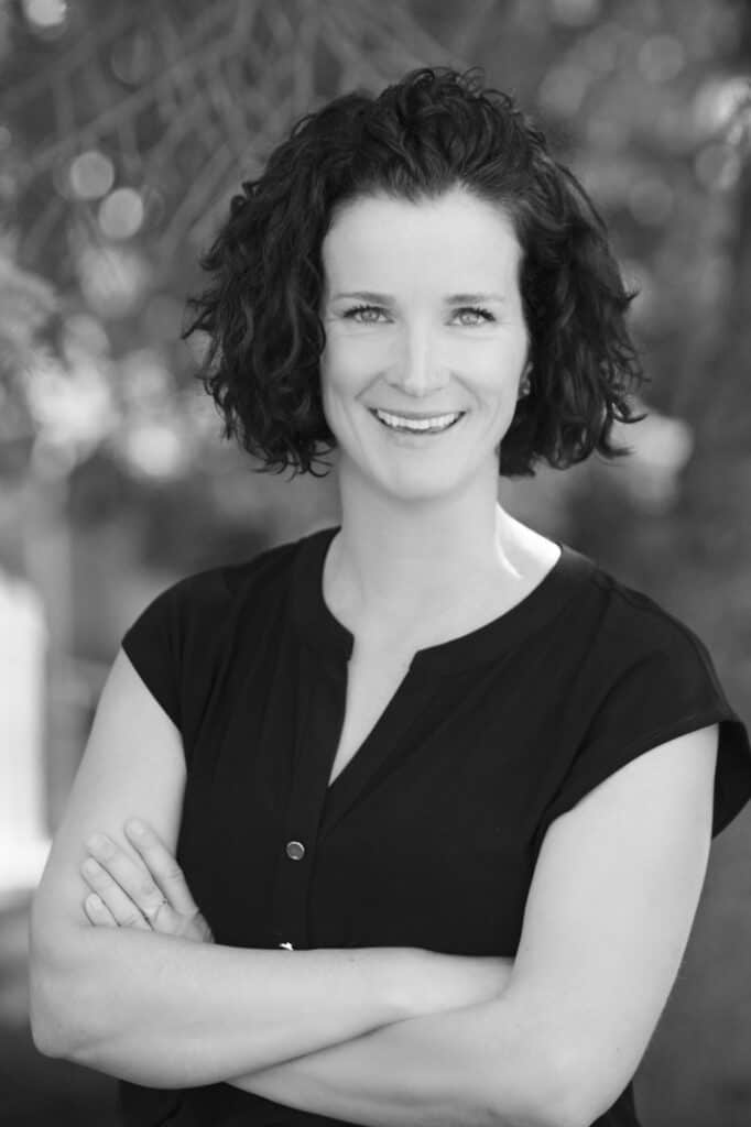 A woman with natural curly dark hair smiles warmly at the camera, standing outdoors with her arms crossed. She is wearing a short-sleeved, dark-colored blouse and the background of blurred trees gives a professional touch. The image is in black and white. - Jodi O Photography