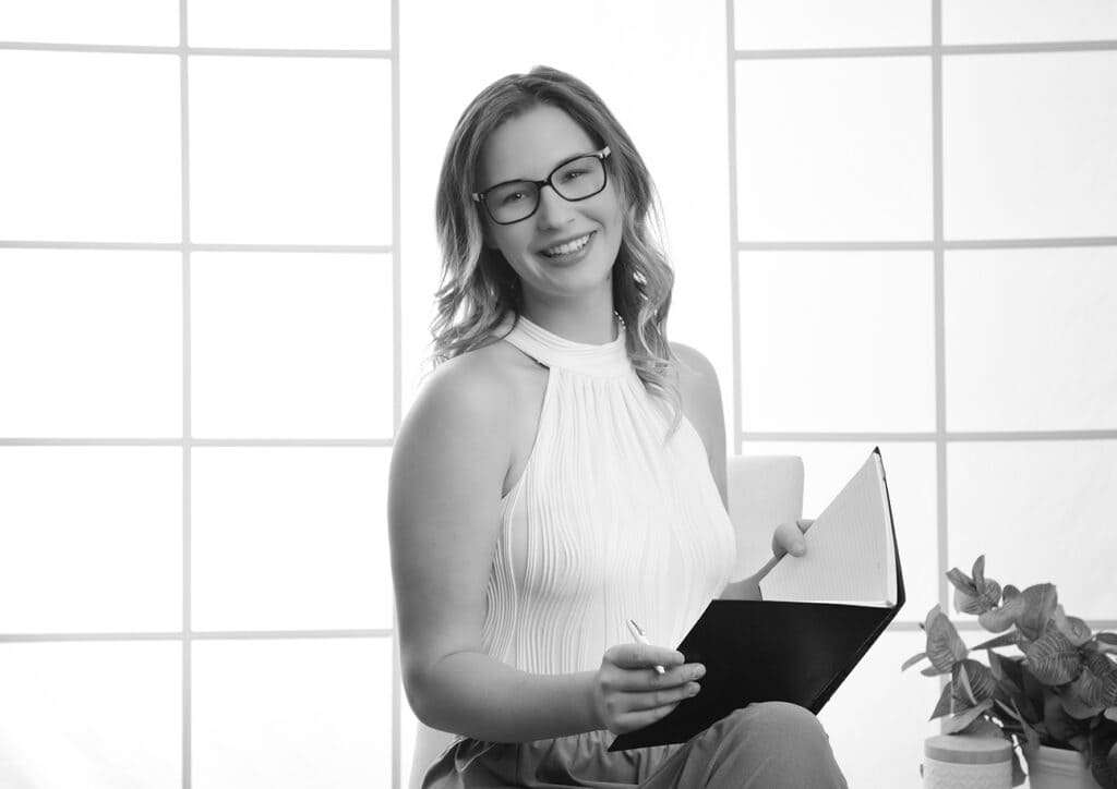 A woman with glasses smiles while holding a pen and an open notebook. Presented in black and white, this modern business image features her by large window panels in a sleeveless top, with a potted plant beside her. - Jodi O Photography